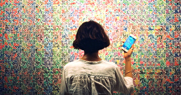 person with short hair standing in front of and facing a multi-colored wall with their phone out ready to record