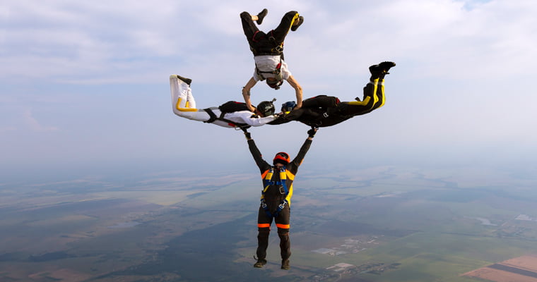 Four people free falling while skydiving. The one on the bottom of the screen has a straight body with arms extending to the two people in the middle. The two people in the middle have their stomachs facing the ground and are mirroring each other. Their arms are bracing each others shoulders with their legs bent up at the knees. The fourth person is at the top with their arms extended slightly bent to the two middle people and pressing down on their upper backs. the person's body is vertical with the knees bent backwards away from the camera.