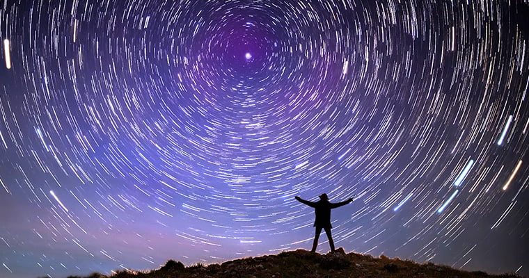 The silhouette of a person standing on top of uneven terrain with arms extended to the sides and looking up at the sky. The sky has a purple to blue to grey gradient that focuses on a center point with the brightest star and swirling stars circling around that point.